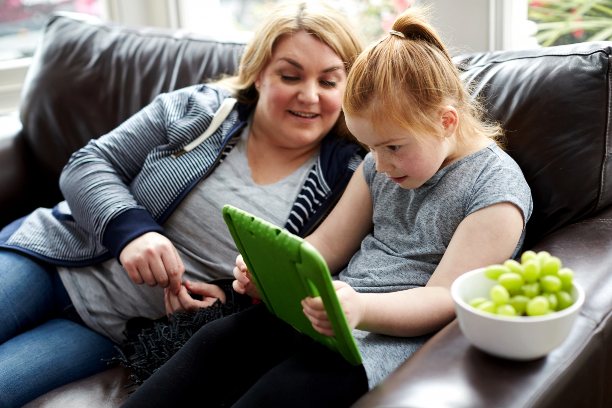 Mother helping daughter use iPad on sofa