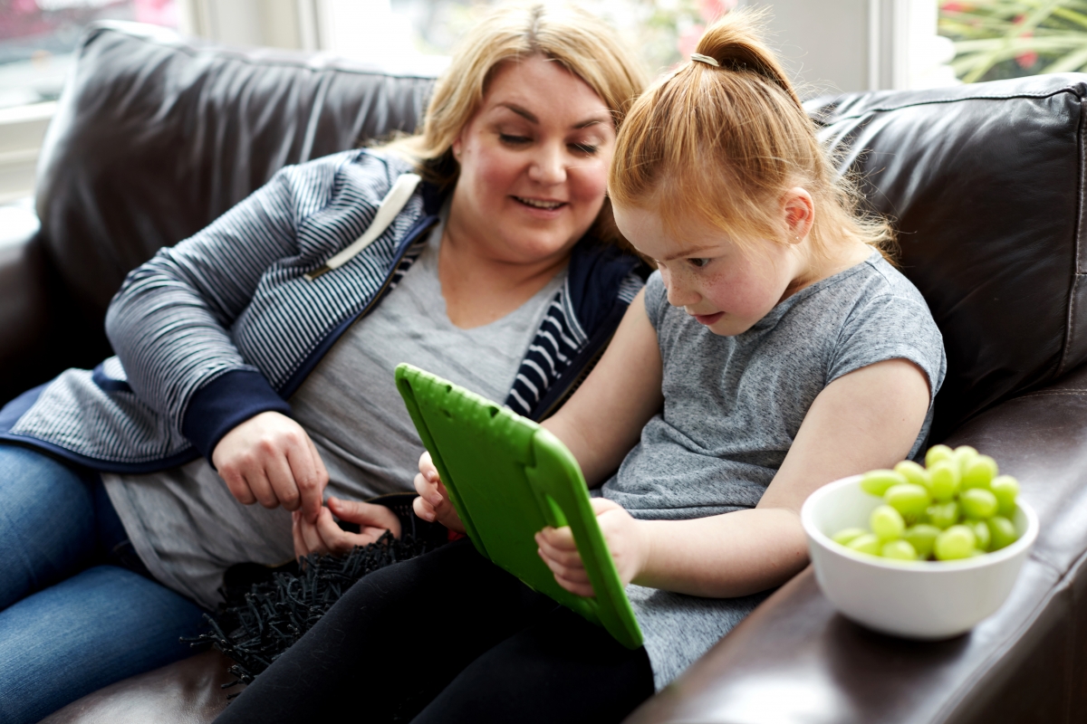 Mother helping daughter use iPad on sofa 